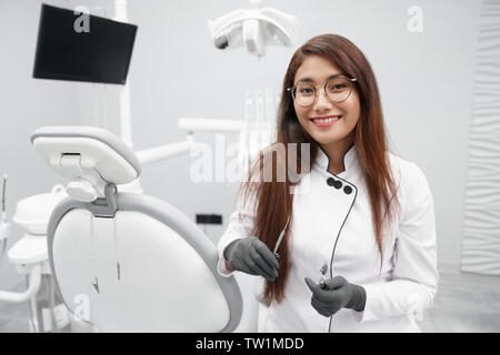 Vue avant du maintien professionnel femme dentiste outils dentaires, looking at camera and smiling in clinique. Pretty doctor posing tout en travaillant en bureau de dentiste. Concept d'emploi et de la stomatologie. Banque D'Images