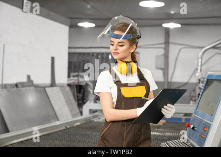 Vue avant du female worker metal factory en processus d'écriture des données. Jolie femme en uniforme et masque de protection, debout près de l'ordinateur et le contrôle de processus cnc et le coupage plasma. Banque D'Images