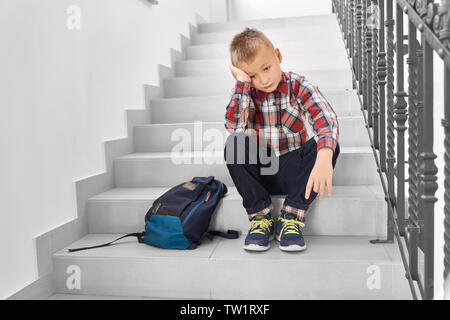 Vue avant du triste garçon blond assis dans les escaliers dans le couloir de l'école et à la main la tête penchée. Peu déçu élève à réfléchir sur son comportement sur lit de leçon. Concept de l'étude. Banque D'Images