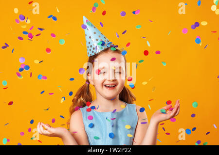 Petite fille joyeuse fête anniversaire. L'enfant est debout avec les yeux fermés dans la pluie de confettis. Closeup portrait sur fond jaune. Banque D'Images