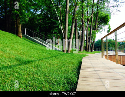Sentier de marche en bois avec garde-corps le long de l'étang dans le parc bien soigné, pelouse verte sur la pente, escalier en béton Banque D'Images