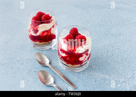 Dessert framboise avec des framboises fraîches, sauce, crème fouettée et de biscuits écrasés dans les verres et les cuillères sur fond de béton bleu with copy space Banque D'Images