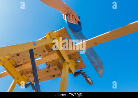 Près de POV shot ci-dessous de ses mains la fin de scier un morceau de bois, qui est fixé dans un portable workbench, à l'extérieur contre un ciel bleu. Banque D'Images