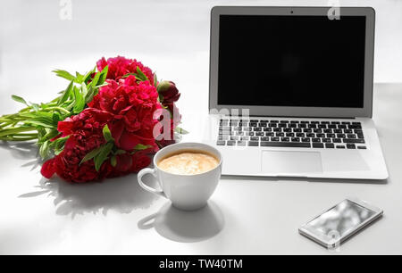 Bouquet de belles pivoines, tasse de café, téléphone mobile et ordinateur sur table lumineuse Banque D'Images