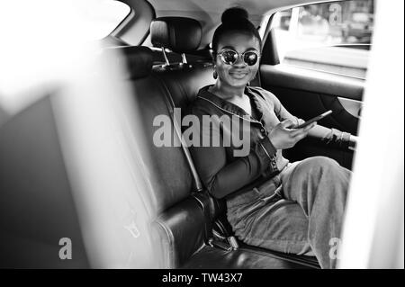 L'entreprise riche femme africaine à siéger à lunettes voiture suv avec des sièges en cuir noir. Téléphone mobile à la main. Banque D'Images