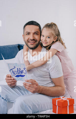 Adorable daughter hugging heureux papa holding pères jours carte de souhaits tout en restant assis près de boîte-cadeau Banque D'Images