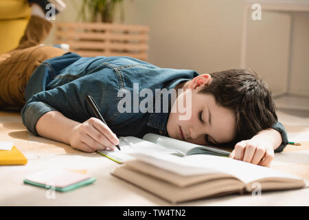 Fatigué garçon couché sur le plancher et l'écriture de copie de livre tout en faisant à la maison schoollwork Banque D'Images