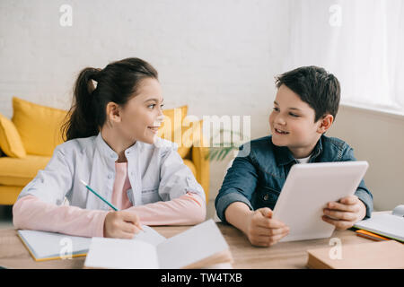 Smiling boy using digital tablet while sitting at desk près de sœur écrit dans l'ordinateur portable Banque D'Images