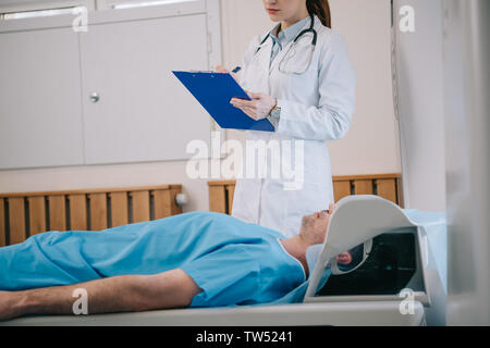 Portrait de jeune radiologue writing on clipboard while standing près de patient allongé sur le lit du scanner IRM Banque D'Images