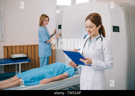 Portrait of smiling radiologue writing on clipboard assistant scanner tout en exploitation près de patient lying on bed scanner Banque D'Images