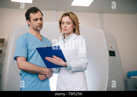 Deux radiologues standing looking au moment du diagnostic sur le presse-papiers en se tenant debout près de tomodensitomètre Banque D'Images