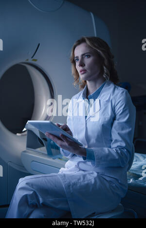 Radiologue réfléchie holding digital tablet with x-ray alors que le diagnostic assis près de tomodensitomètre et à l'écart Banque D'Images
