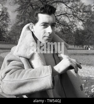 Années 1960, historiques, les jeunes "l'homme à propos de town' debout dans un parc, dans les modes du jour, porter un costume de pull en laine, un cravat, une épaisse veste sheepsking et fumer la cigarette, England, UK. Banque D'Images
