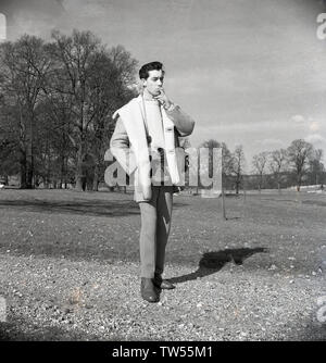 Années 1960, historiques, photographe...un jeune homme à propos de town' avec son appareil photo, debout sur un sentier de gravier à l'extérieur dans un parc à la mode du jour, porter un costume, un pull en laine et une épaisse cravate veste en peau de mouton et de fumer la cigarette, England, UK. Banque D'Images