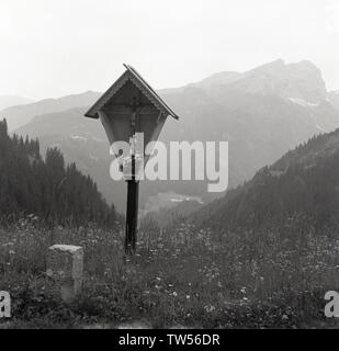 Années 1950, historique, d'un vase de fleurs Daisy par une petite statue d'un prêtre à l'enfant debout sous une sculpture de Jésus sur la croix, à l'intérieur d'un petit temple en bois se tenait sur la colline dans l'Italian Dolomites, Italie. Banque D'Images