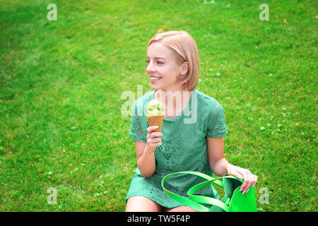 Young happy woman in green hippie robe dentelle holding ice cream while sitting on grass in park Banque D'Images