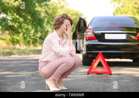 Contrarié woman sitting on road, près de sa voiture cassée Banque D'Images