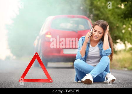 Jeune femme en colère avec cell phone assis sur route près de la voiture cassée Banque D'Images
