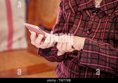 Vieille Femme Asiatique assis et regarder à travers quelque chose sur smartphone moderne, faisant la liaison avec d'autres personnes à la maison, un salon de la technologie, Close up Banque D'Images