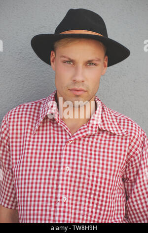 Un modèle masculin blond portant une chemise à carreaux rouge de l'Ouest, et un chapeau fedora posé contre un mur debout à l'extérieur. Un portrait en gros plan. Banque D'Images