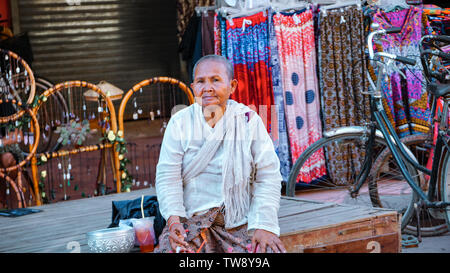 Phnom Penh, Cambodge - Août 2016 : Une vieille femme cambodgienne assis sur rue en face de boutiques décorées avec des chaises et des robes. Banque D'Images