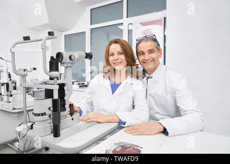 Vue de face de deux oculists avec travail à la lampe à fente ensemble en magasin d'optique. Deux yeux doctors sitting at table, looking at camera, souriant et posant en clinique. Concept d'ophtalmologie et de la santé. Banque D'Images