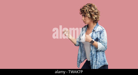 Vue côté profil portrait de jeune femme sérieuse avec curly hairstyle in casual chemise bleue avec les poings boxe permanent, à l'avant, prêt à l'atta Banque D'Images