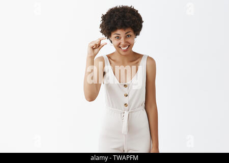 D'humour et d'émotion esthétique african american woman with curly hairstyle en combinaisons blanches montrant quelque chose de minuscule, façonner petit article avec les doigts Banque D'Images