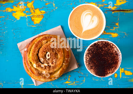 Deux cappuccinos et cannelle rouleau à pâtisserie isolé sur l'épluchage et craquelé bleu et jaune au-dessus de bois peint. Banque D'Images