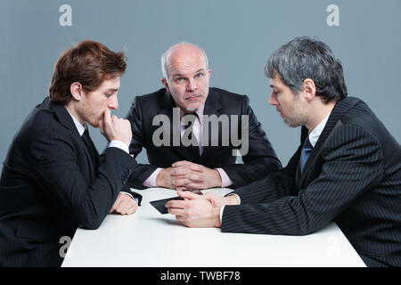 Trois hommes d'avoir une discussion sérieuse au cours d'une réunion assis autour d'une table de bureau avec l'homme plus âgé à la recherche jusqu'à l'appareil photo de manière réfléchie Banque D'Images