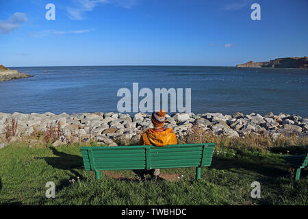 Une femme portant des vêtements d'hiver se trouve sur un banc vert, à la SAE Nord à Runswick Bay, North Yorkshire, UK. Banque D'Images