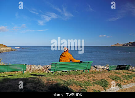 Une femme portant des vêtements d'hiver se trouve sur un banc vert, à la SAE Nord à Runswick Bay, North Yorkshire, UK. Banque D'Images