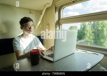 Un adolescent garçon sur un train est assis à une table avec un ordinateur ouvert Banque D'Images