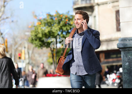 Jeune homme urbain on smart phone fonctionnant en parlant de la rue sur smartphone smiling portant veste et sac pour ordinateur portable en cuir sur le Passeig de Gracia, Barcelone, Catalogne, Espagne. Banque D'Images