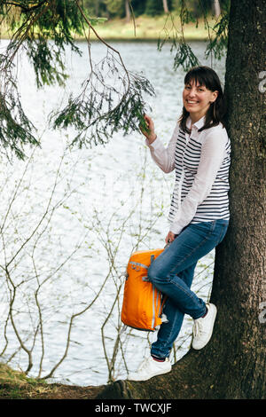 Jeune fille de tourisme avec sac à dos orange dans les mains, debout près de pour le grand arbre sur la rive du lac de montagne entouré par la forêt. Banque D'Images