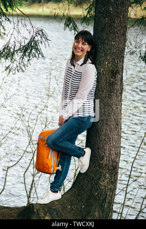 Les jeunes touristes smiling girl avec sac à dos orange dans les mains, debout près de pour le grand arbre sur la rive du lac de montagne entouré par la forêt. Banque D'Images