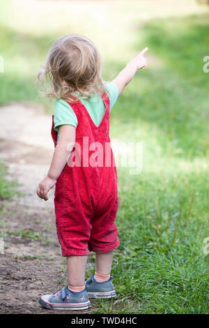 Petit enfant en vert tee-shirl et pantalon rouge debout dos et dirigés à l'écart avec l'index sur fond de plein air d'été Banque D'Images