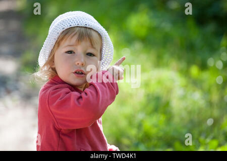 Petit enfant dans la veste rouge et blanc hat pointe vers le haut à l'index vue rapprochée sur fond extérieur vert Banque D'Images