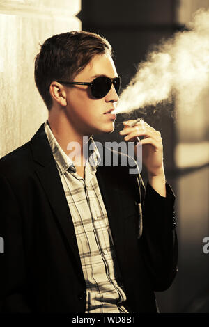 Jeune homme à lunettes fumer une cigarette on city street fashion élégant modèle masculin vêtu de noir veste de costume Banque D'Images