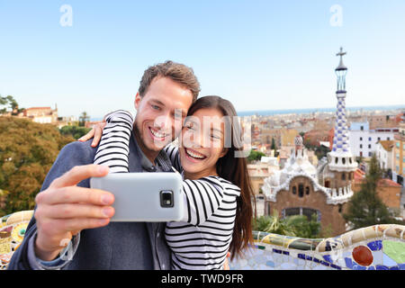 Billet couple heureux de parler de selfies smartphone dans le parc Guell, Barcelone, Espagne. Beau jeune couple multiracial en tenant l'appareil photo avec smart phone smiling dans l'amour. Banque D'Images