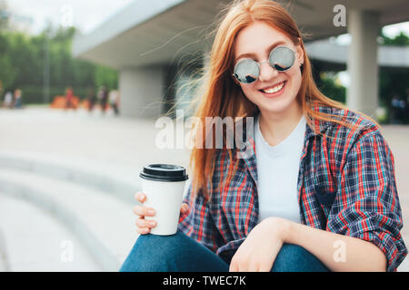 Jolie rousse smiling girl in lunettes rondes avec tasse de café pour aller dans les mains dans les tenues sur rue dans la ville Banque D'Images