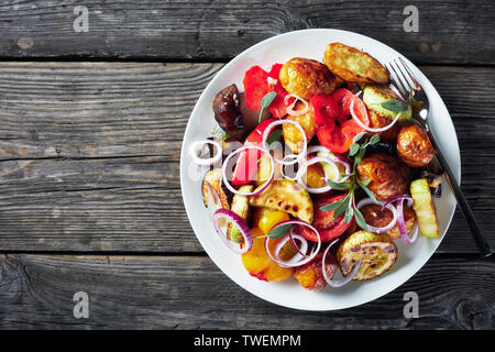 Rôtie au four, légumes chauds - pommes de terre, aubergines, courgettes, tomates, poivrons sur une assiette blanche sur une table en bois gris, horizonta Banque D'Images