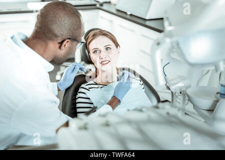 Femme qui a problème. Un médecin travaillant à la peau sombre avec sa jolie patiente transportant des outils de métal dans les mains couvertes Banque D'Images