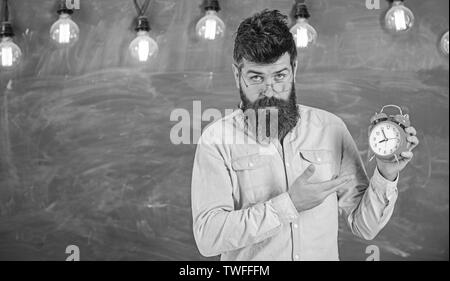 Enseignant dans les lunettes est titulaire d'un réveil. Concept de discipline. Homme avec barbe et moustache sur la lettre, se tenir dans la salle de classe. Hipster barbu est titulaire d'réveil, tableau sur toile de fond, l'espace de copie. Banque D'Images