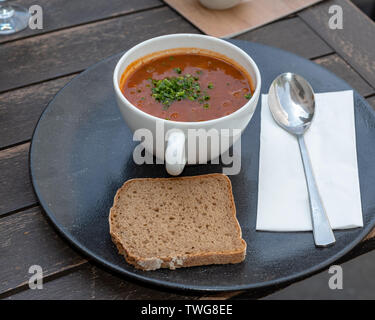 Une plaque noire avec Goulash garnie d'oignons de printemps, servi dans une tasse de thé, un morceau de pain, une cuillère et une serviette sur une table en bois à l'extérieur setti Banque D'Images