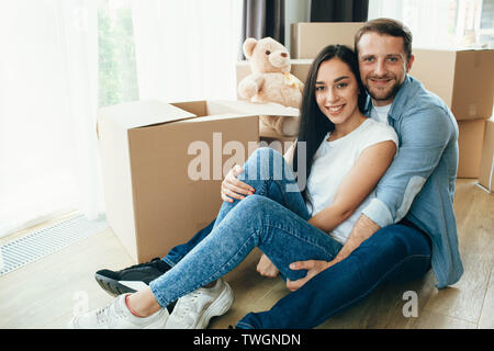 Heureux Couple hugging and sitting on floor dans leur nouvelle maison avec de nombreux cartons on background Banque D'Images