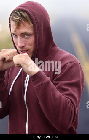 Boxe boxeur de remise en forme à l'extérieur sur la route in hoodie. Homme de perforation et de frapper à la recherche à l'appareil photo. Adapter la forme physique dans la transpiration boxer noir à l'extérieur. Homme de race blanche dans son modèle de remise en forme 20s. Banque D'Images