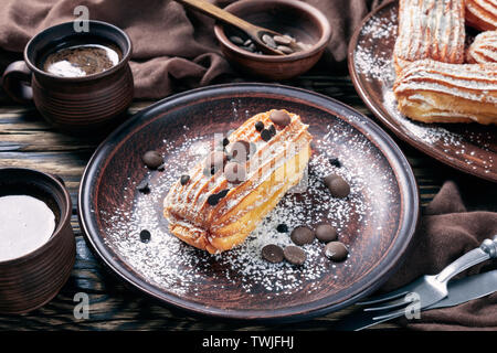 De délicieux éclairs Maison saupoudré de sucre en poudre et de pastilles de chocolat sur une plaque de faïence sur une vieille table rustique avec tasse de café, ho Banque D'Images