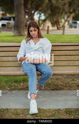 Une jeune fille est assise sur un banc de parc en utilisant son téléphone. Banque D'Images