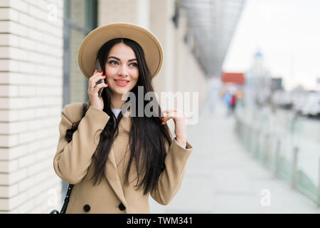 Beau portrait femme aux longs cheveux noirs de parler par téléphone avec l'ami en se tenant sur le sunny street, brunette wearing manteau beige, à l'écart, Banque D'Images